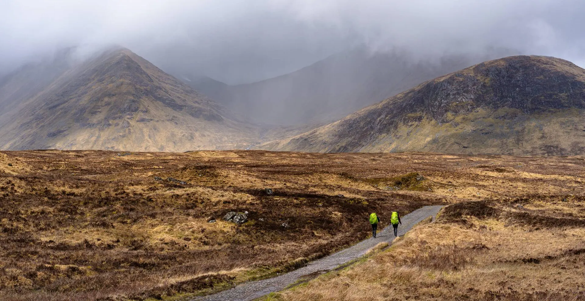 two persons with green backpacks hiking in misty moorland