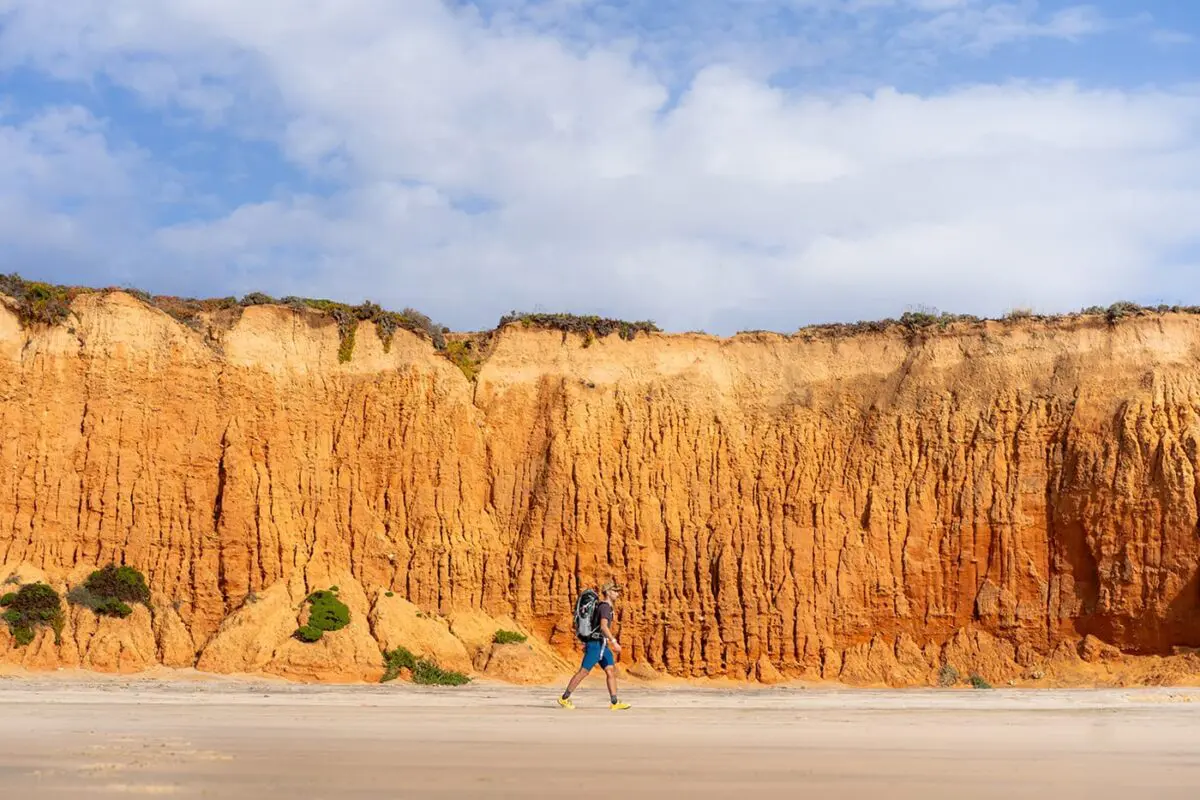 man hiking on beach with orange cliffs behind him