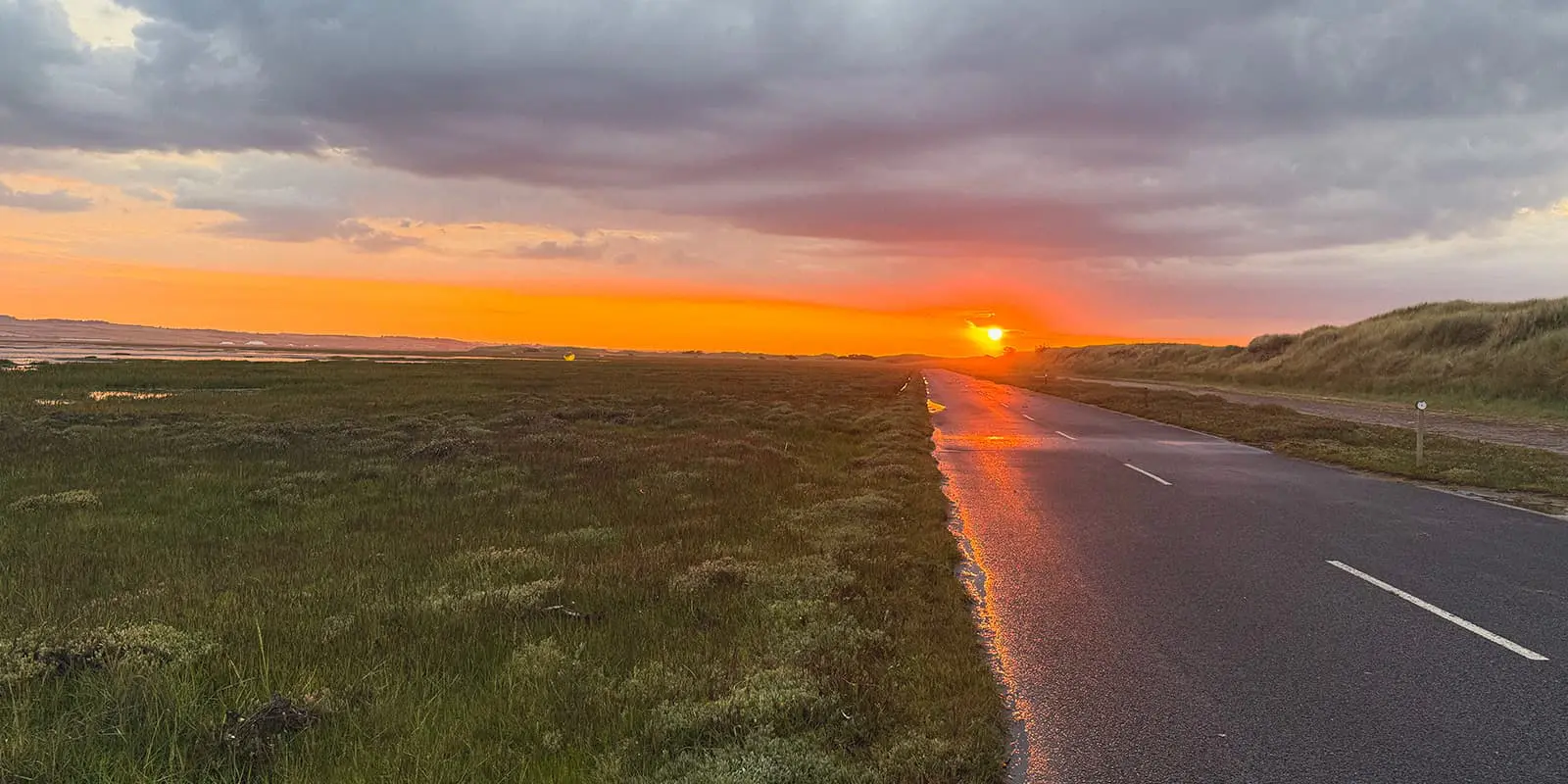 empty road next to green fields and body of water with sunset in the background