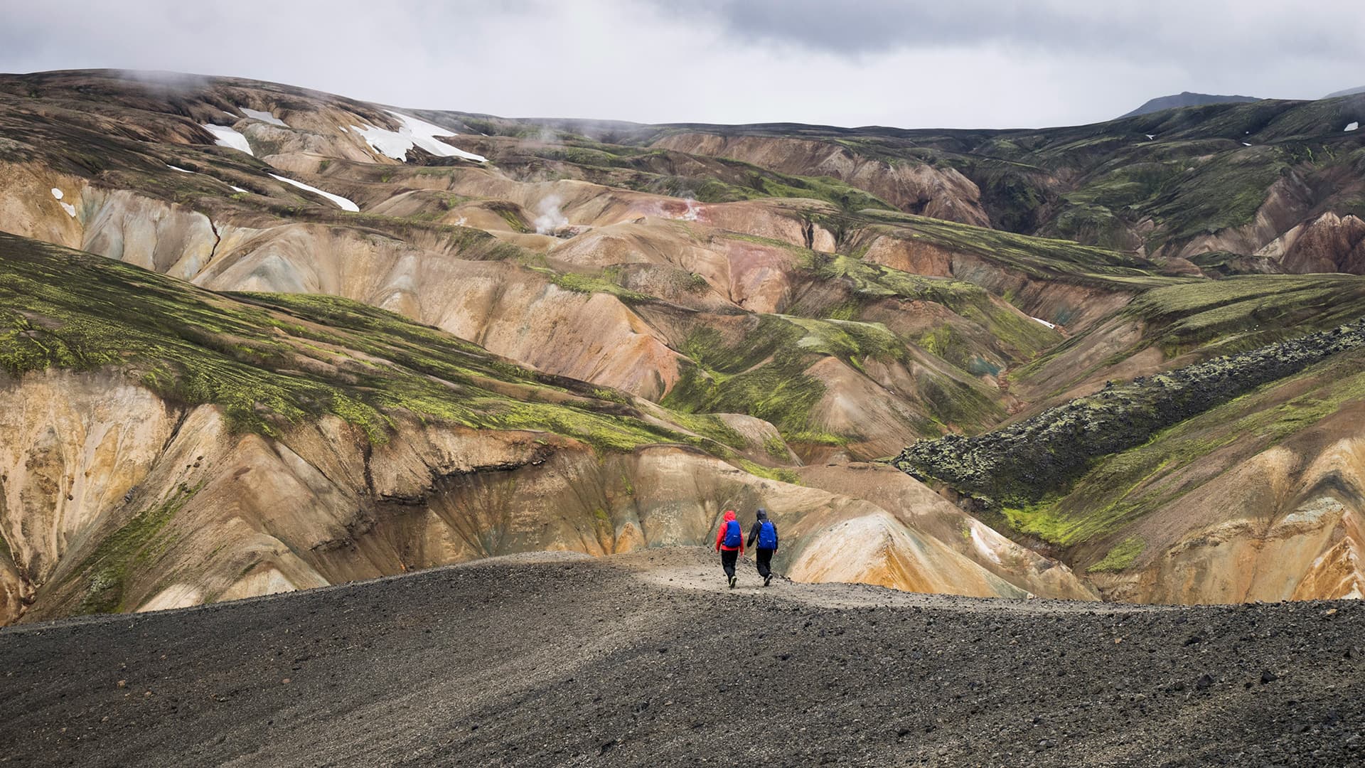 Two hikers walking in distance on black sand with colored mountains in the background