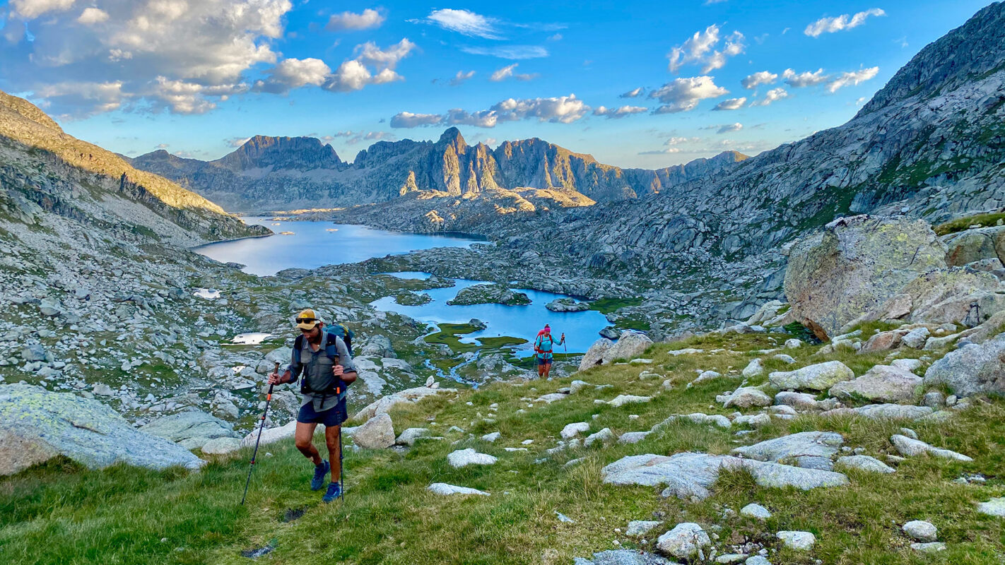 Hiker walking on mountain trail with small lake and mountains in the background during sunrise