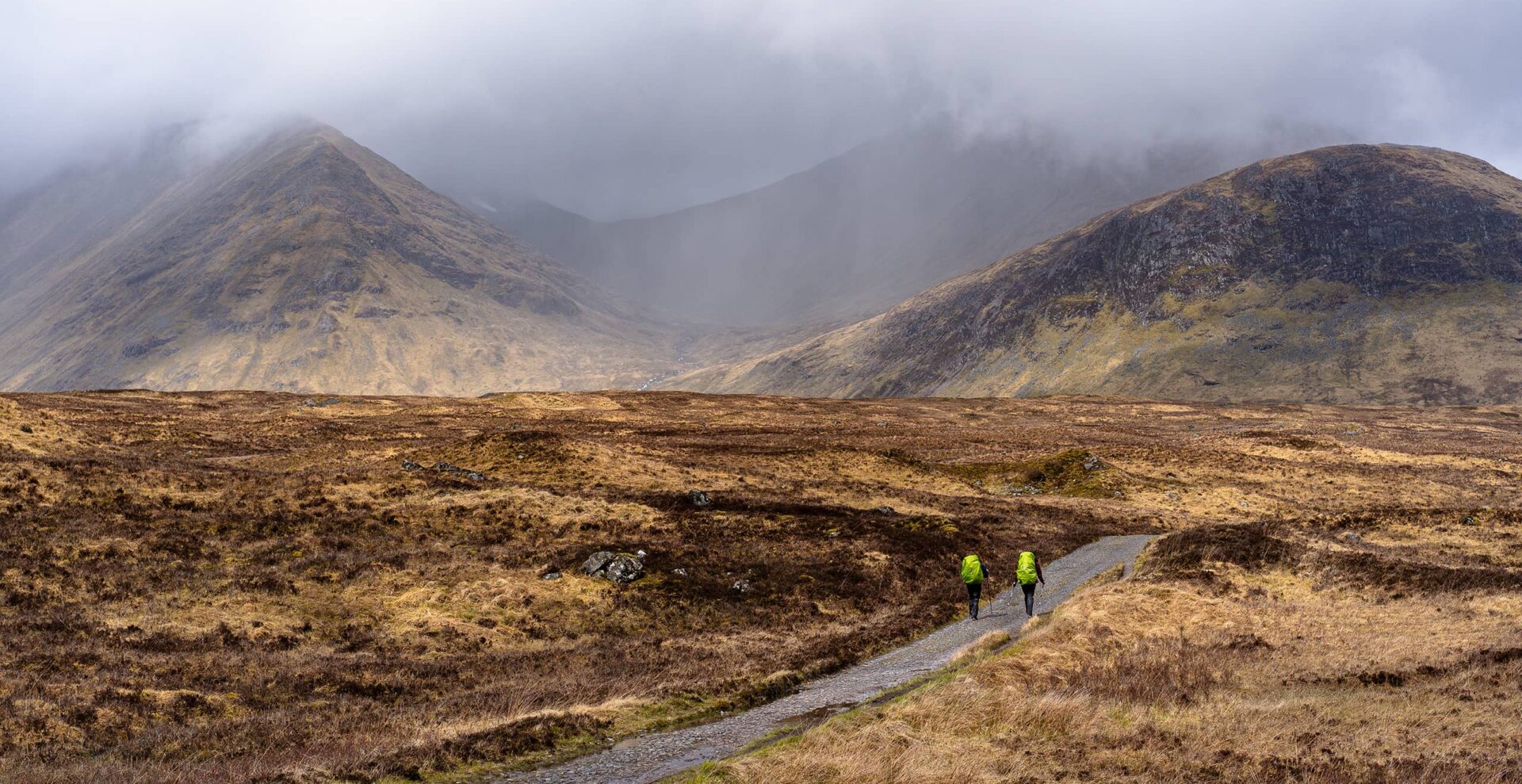 two persons with green backpacks hiking in misty moorland