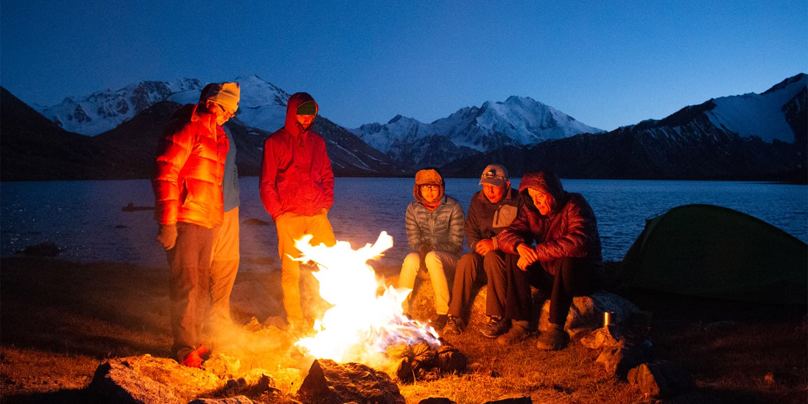 group of people sitting by the fire near mountain lake